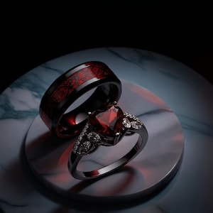 Op de afbeelding: Twee ringen: een zwarte band met een rood drakenpatroon en een zwarte ring met een hartvormige rode edelsteen en kleine witte edelstenen. De ringen worden gepresenteerd op een marmeren oppervlak.