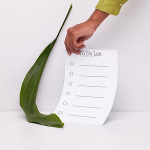 May include: A white To Do List with checkboxes is held by a hand, next to a large green leaf. The text "To Do List" is at the top of the page. The background is white.