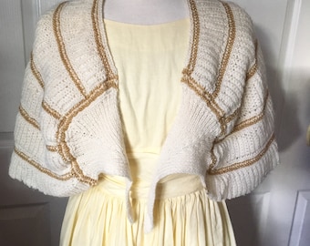 Puede incluir: Una estola de punto blanco con rayas doradas. La estola está drapeada sobre un vestido amarillo.