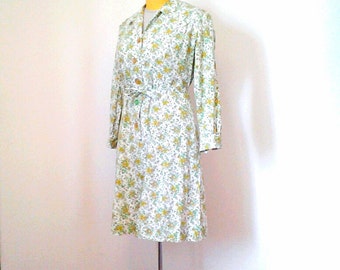 Puede incluir: Un vestido blanco con estampado floral en tonos amarillos y verdes. El vestido tiene manga larga, cuello de camisa y cinturón en la cintura.