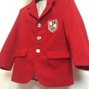 May include: A bright red blazer with silver buttons and a crest on the chest. The blazer has two front pockets and a notched lapel. The crest is white with a design of crowns and other symbols. The blazer is hanging on a black hanger.