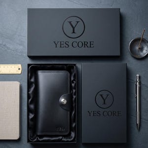 Op de afbeelding: Een zwarte lederen portemonnee met een zilveren drukknoopsluiting, gepresenteerd in een zwarte geschenkdoos met de tekst YES CORE en een Y-logo. De set bevat een metalen liniaal, een klein notitieboekje, een metalen pen en een wierookhouder.