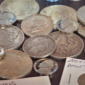 May include: A collection of silver coins, including a Peace dollar, a Walking Liberty half dollar, and several other coins. The coins are arranged on a brown surface. The text "2004 P MS65 Peace" is visible on a white piece of paper.
