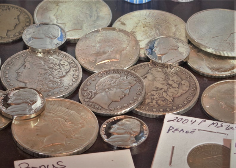 May include: A collection of silver coins, including a Peace dollar, a Walking Liberty half dollar, and several other coins. The coins are arranged on a brown surface. The text "2004 P MS65 Peace" is visible on a white piece of paper.