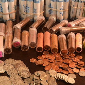 May include: A collection of rolled US coins in plastic and paper tubes. The coins are arranged on a wooden surface with a pile of loose silver coins in the foreground. The tubes are labeled with the coin denomination and year.