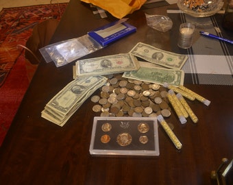 May include: A wooden table with a collection of US currency, coins, and gold flakes in small glass tubes. There is a clear plastic bag with a few coins inside, a blue box with white lettering, and a small black box with a collection of coins inside.