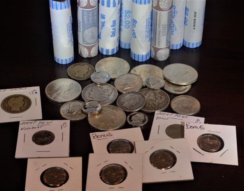 May include: A collection of silver coins on a dark surface, with some of the coins arranged on small white cards with handwritten labels. The labels include "Bonus", "Peace", "Keelboat", and "Bonus".