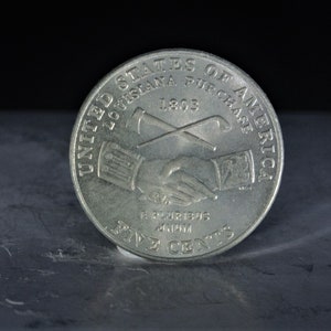 May include: A silver United States five-cent coin commemorating the Louisiana Purchase. The coin features a handshake between two figures, an axe and a shovel crossed behind them, and the year 1803. The inscription reads "United States of America, Louisiana Purchase 1803, E Pluribus Unum, Five Cents."
