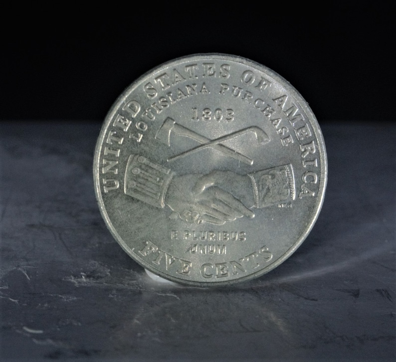 May include: A silver United States five-cent coin commemorating the Louisiana Purchase. The coin features a handshake between two figures, an axe and a shovel crossed behind them, and the year 1803. The inscription reads "United States of America, Louisiana Purchase 1803, E Pluribus Unum, Five Cents."