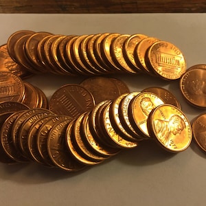 May include: A collection of copper-coloured US pennies, some stacked in piles and others scattered on a white surface. The pennies feature the Lincoln Memorial and the words "One Cent" and "United States of America".