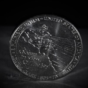 May include: A silver United States quarter dollar coin with the words "E Pluribus Unum" and "United States of America" around the edge. The coin features a depiction of the Lewis and Clark Expedition with the words "Lewis & Clark 1805-1806" and "Great Western View" on the coin.