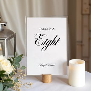 以下が含まれることがあります： エレガントな黒い筆記体で「Eight」と書かれた透明なアクリル製のテーブルナンバーサイン。サインには上部に「Table No.」、下部に「Henry & Eleanor」という文字が含まれています。小さな木製のベースの上に置かれ、近くには火の灯ったキャンドルと花の飾りがあります。