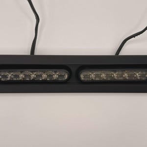May include: A black rectangular light fixture with two rows of six clear LED lights. The fixture has a black frame and two black wires extending from the top.