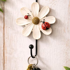 May include: A daisy-shaped key holder with two ladybug decorations, hanging on a white wall. The key holder has a black metal hook and a ring holding keys and a car remote. The logo "HJEM HAVEN BY ELISE" is in the lower left corner.