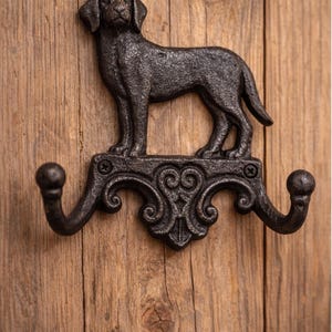 May include: Black cast iron dog-shaped wall hook. The hook features a detailed Labrador Retriever design above two curved hooks with decorative scrollwork. Mounted on a wooden background.