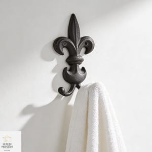 Könnte beinhalten: Ein schwarzer Wandhaken in Form einer Lilie mit einem weißen Handtuch. Der Haken ist aus Metall und hat ein dekoratives Design. Das Handtuch ist aus weichem Material. Der Haken ist an einer weißen Wand befestigt.