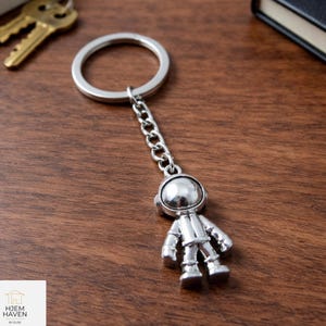 May include: A silver astronaut keyring with a chain and ring. The astronaut figure has a helmet, suit, and boots. The keyring is shown on a wooden surface, with keys and a small notebook in the background.