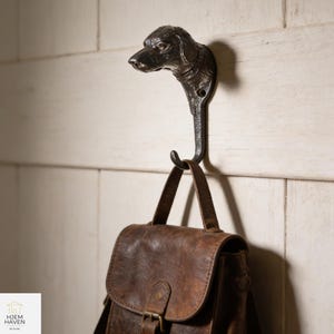 Könnte beinhalten: Ein dunkler Metallhaken in Form eines Hundekopfes, montiert an einer weißen Holzwand. Eine braune Ledertasche hängt an dem Haken. Der Hundekopf ist mit einem Halsband und detaillierten Merkmalen versehen. Ein dekorativer und praktischer Artikel.
