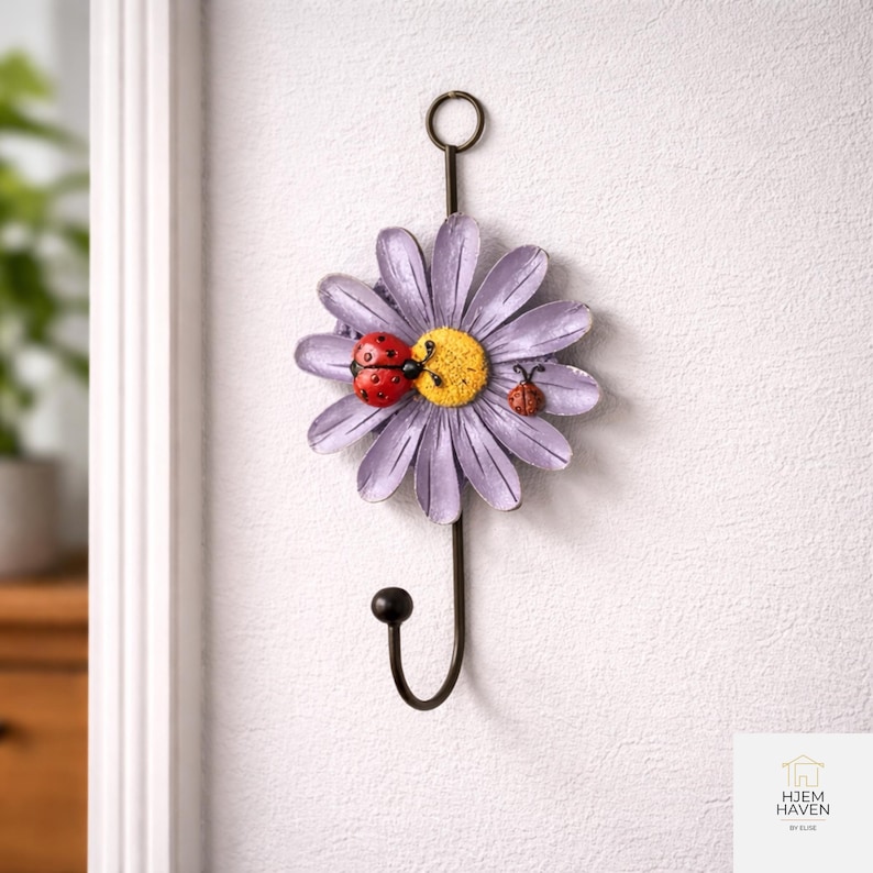 Op de afbeelding: Een decoratieve metalen wandhaak in de vorm van een madeliefje met paarse bloemblaadjes en een gele kern. Twee rode lieveheersbeestjes zitten op de bloem. De zwarte haak heeft een lus aan de bovenkant om op te hangen.