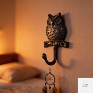 Könnte beinhalten: Ein dunkelgrauer Metall-Eulen-Haken an der Wand, mit einem dekorativen Traumfänger, der am Haken hängt. Die Eule sitzt auf einem Ast, und der Haken ist gebogen. Die Wand ist hellbeige.