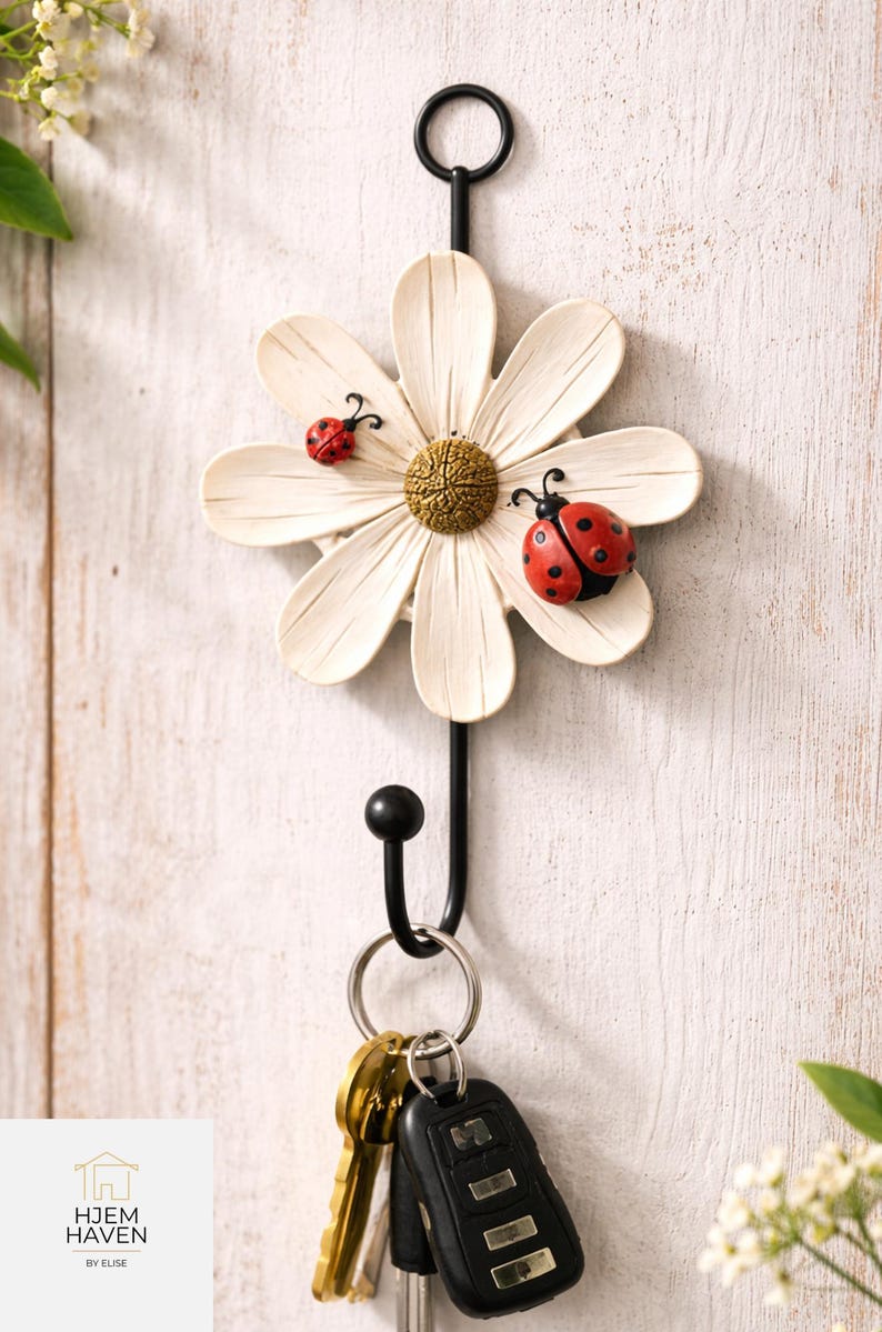 Op de afbeelding: Een decoratieve sleutelhouder voor aan de muur in de vorm van een witte madelief met twee lieveheersbeestjes. De sleutelhouder heeft een zwarte metalen haak en een ring met sleutels en een afstandsbediening voor de auto. Het logo "HJEM HAVEN BY ELISE" staat linksonder.