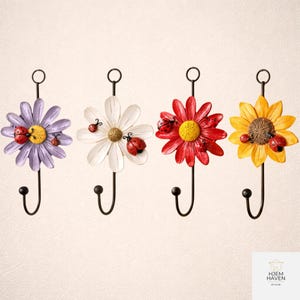Op de afbeelding: Vier decoratieve wandhaken met bloemmotieven in paars, wit, rood en geel. Elke haak heeft een zwarte metalen steel en een lieveheersbeestje accent. De haken zijn ontworpen om items op te hangen.