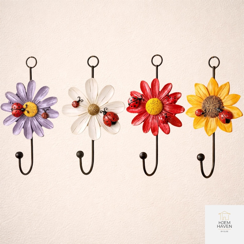 Op de afbeelding: Vier decoratieve wandhaken met bloemmotieven in paars, wit, rood en geel. Elke haak heeft een zwarte metalen steel en een lieveheersbeestje accent. De haken zijn ontworpen om items op te hangen.