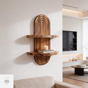 May include: A wooden wall shelf with two circular shelves and a decorative arched top. A lit candle and two books rest on the lower shelf. The shelf is made of light brown wood and is mounted on a white wall.