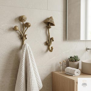 May include: Two brass mushroom-shaped wall hooks. One hook features a cluster of three mushroom heads, and the other has a single mushroom cap design. A white waffle-weave towel hangs from the first hook. The hooks are mounted on a light-colored tiled wall.