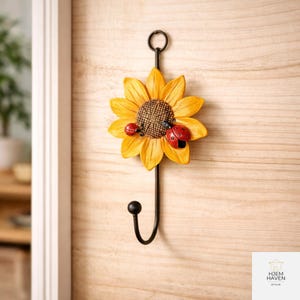 Op de afbeelding: Een decoratieve wandhaak met een gele zonnebloem met een bruin hart en twee rode lieveheersbeestjes. De haak is gemaakt van zwart metaal en heeft een lus aan de bovenkant om op te hangen.