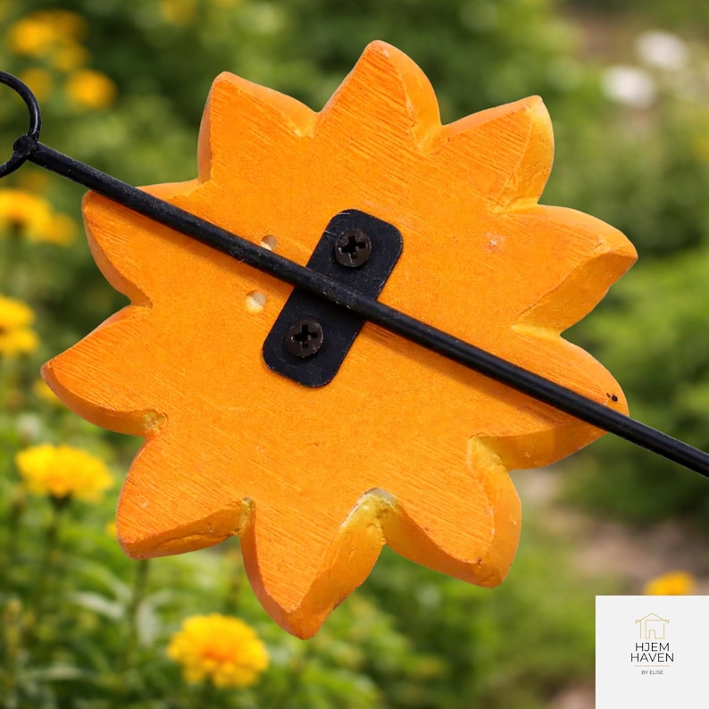 Op de afbeelding: Close-up van een oranje, houten, bloemvormig tuinornament. Het ornament heeft een zwarte metalen staaf aan de achterkant bevestigd met twee schroeven. De achtergrond is wazig en toont groene bladeren en gele bloemen.