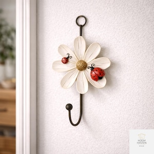 Op de afbeelding: Een decoratieve wandhaak in de vorm van een witte madelief met een gouden hart en twee rode lieveheersbeestjes. De haak is gemaakt van donker metaal en is ontworpen om aan de muur te hangen. Het ontwerp is ge&iuml;nspireerd op de natuur.