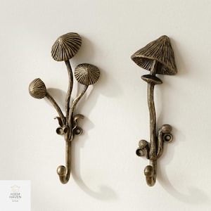 May include: Two bronze-colored mushroom-shaped wall hooks. One hook features three mushroom caps, while the other has a single, larger cap. Both have a hook at the bottom and decorative stems. The hooks are mounted on a white wall.