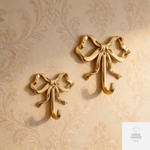 May include: Two gold-coloured bow-shaped wall hooks. The hooks are made of metal with a glossy finish. The bows have detailed folds and a hook at the bottom. The hooks are mounted on a beige wall with a floral pattern.