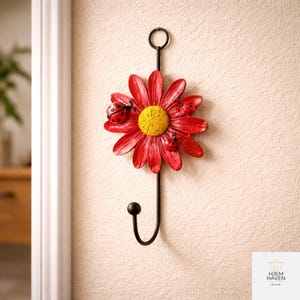 Op de afbeelding: Een decoratieve wandhaak met een rode madelief met een gele kern en drie lieveheersbeestjes. De haak is zwart en heeft een lus aan de bovenkant om op te hangen. De madelief heeft een diameter van ongeveer 20 cm.