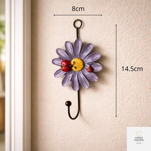 Op de afbeelding: Een decoratieve wandhaak in de vorm van een paarse madelief met een gele kern en twee rode lieveheersbeestjes. De zwarte haak is 14,5 cm lang en 8 cm breed. Het madeliefontwerp voegt een vleugje door de natuur ge&iuml;nspireerde charme toe.
