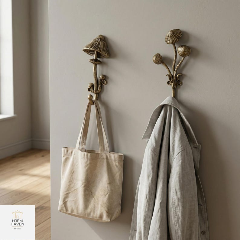 May include: Two bronze-colored mushroom-shaped wall hooks. One hook holds a beige tote bag, and the other holds a light gray jacket. The hooks are mounted on a light gray wall. The lower left corner has a white square with the text "HJEM HAVEN BY ELISE".
