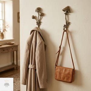 May include: Two bronze mushroom-shaped wall hooks with a beige coat and a brown leather satchel hanging from them. The hooks are mounted on a cream-colored wall. The coat is a long, belted style. The satchel has a flap closure.