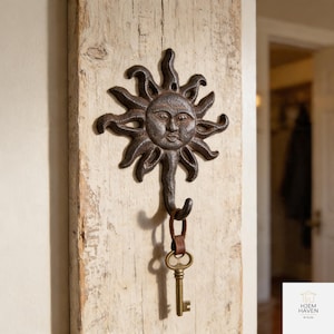 May include: A rustic, cast iron sun-shaped wall hook with a key hanging from it. The hook is mounted on a weathered, light-colored wooden plank. The sun has a detailed face and radiating rays, with a bronze-colored key attached.