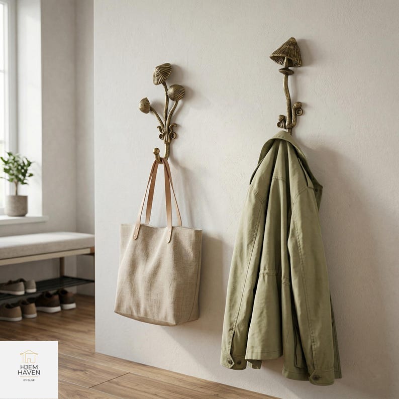 May include: Two decorative mushroom-shaped wall hooks in a bronze finish. One hook holds a beige tote bag with leather handles, and the other holds a green jacket. The hooks are mounted on a light-colored wall.