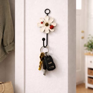 Op de afbeelding: Een decoratieve aan de muur gemonteerde sleutelhouder met een wit madeliefjesontwerp en twee rode lieveheersbeestjes. De zwarte metalen haak houdt een set sleutels vast. De sleutelhouder is aan een witte muur gemonteerd.