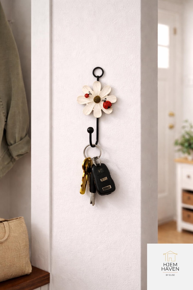 Op de afbeelding: Een decoratieve aan de muur gemonteerde sleutelhouder met een wit madeliefjesontwerp en twee rode lieveheersbeestjes. De zwarte metalen haak houdt een set sleutels vast. De sleutelhouder is aan een witte muur gemonteerd.