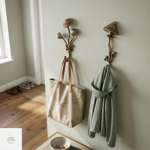 May include: Two decorative brass mushroom-shaped wall hooks. One hook holds a beige tote bag with leather straps, and the other holds a green jacket. The hooks are mounted on a light green wall, creating a whimsical and functional storage solution.