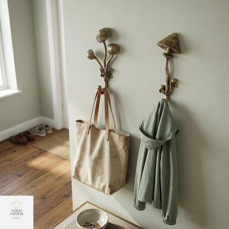 May include: Two decorative brass mushroom-shaped wall hooks. One hook holds a beige tote bag with leather straps, and the other holds a green jacket. The hooks are mounted on a light green wall, creating a whimsical and functional storage solution.