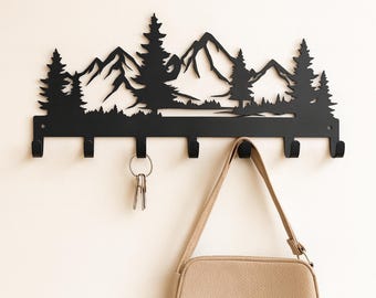 May include: Black metal wall-mounted key holder featuring a mountain and forest silhouette design. The key holder has five hooks and is holding a set of keys and a beige purse. The design includes trees and mountain peaks.