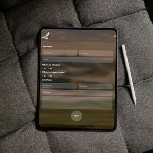 May include: A black tablet displaying an online form with fields for name, email, and RSVP. A white stylus rests beside the tablet on a gray, textured surface. The form includes the text "Your Name", "Email", "Will you be attending?", "Will you be bringing a guest?", "Guest Name", and "RSVP".