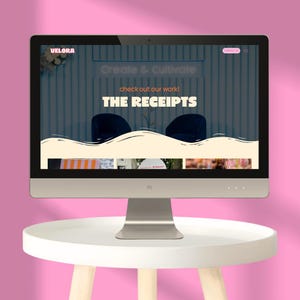 May include: A computer monitor displaying a website with the text Velora, Create & Cultivate, check out our work!, and THE RECEIPTS. The screen features a cream-colored wavy graphic element over a dark background with two dark blue chairs.