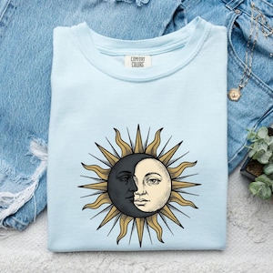 May include: A light blue crew neck t-shirt with a Comfort Colors brand label at the inner neckline. The center of the shirt features a graphic illustration of a sun and moon combined, with gold rays, a dark crescent moon, and a cream-colored face.