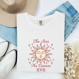 May include: A white crew neck t-shirt featuring a red graphic of a sun with a face, surrounded by small star icons. The text reads The Star The Cosmos XVII. The shirt is displayed with a tan hat, blue denim jeans, and white boots.