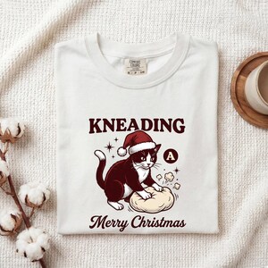 May include: A white t-shirt featuring a graphic of a black and white cat wearing a red Santa hat, kneading dough. The text reads KNEADING above the cat and Merry Christmas below. The cat is positioned next to a small circle containing the letter A.